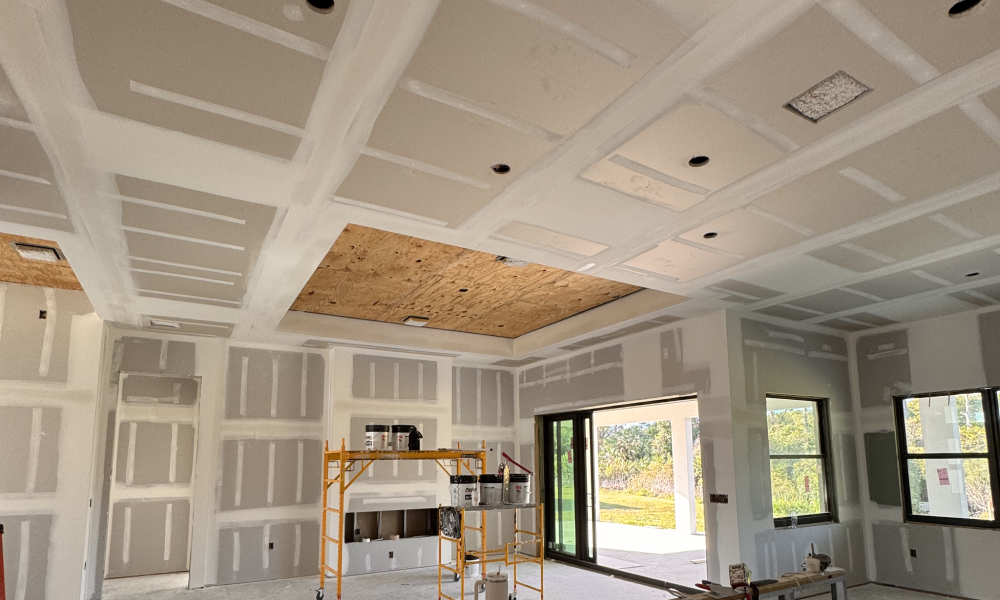 The interior of a room under construction features drywall and a large unfinished ceiling patch, complemented by recessed lighting fixtures. Construction materials and a scaffold with paint cans suggest the presence of a general contractor. Large windows flood the space with natural light.