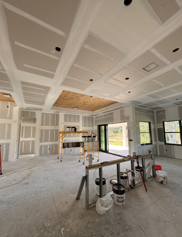 A partially finished room with drywall installation complete on the walls and ceiling. An opening leads to a patio, and a window offers an outdoor view. A scaffold holds tools, while the flooring installation areas are scattered with construction supplies and containers.