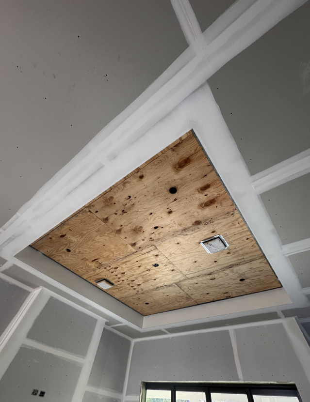A photo of a partially finished ceiling under construction showcases expert drywall installation. The ceiling features a recessed center covered with plywood, two visible light fixtures, and a glimpse of a window—craftsmanship typical of an experienced kitchen remodeler.