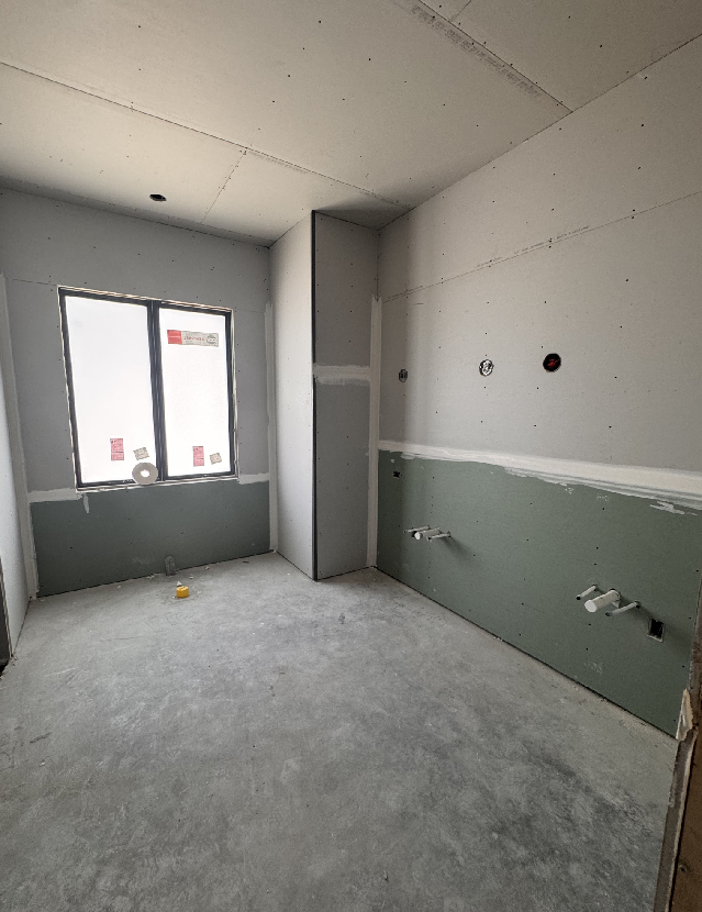 An unfinished interior space with unpainted drywall and a concrete floor awaits transformation by a skilled general contractor. Plumbing fixtures on one wall hint at a future bathroom, while two large windows flood the room with natural light.