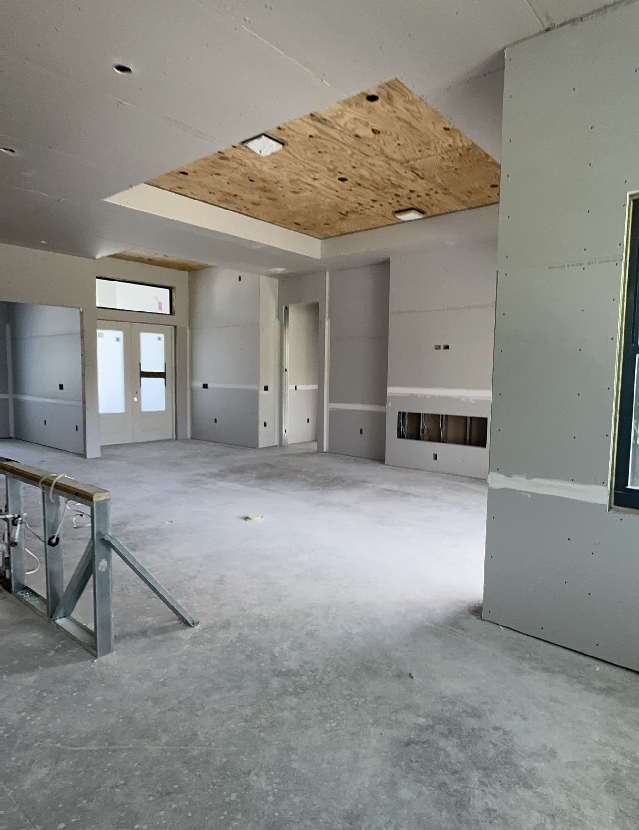 The image captures the interior of a house under construction, highlighting dry wall installation with unfinished walls and ceiling. A light wooden board covers part of the ceiling, while a window graces the right. To the left, scaffolding hints at the skilled work of a general contractor.