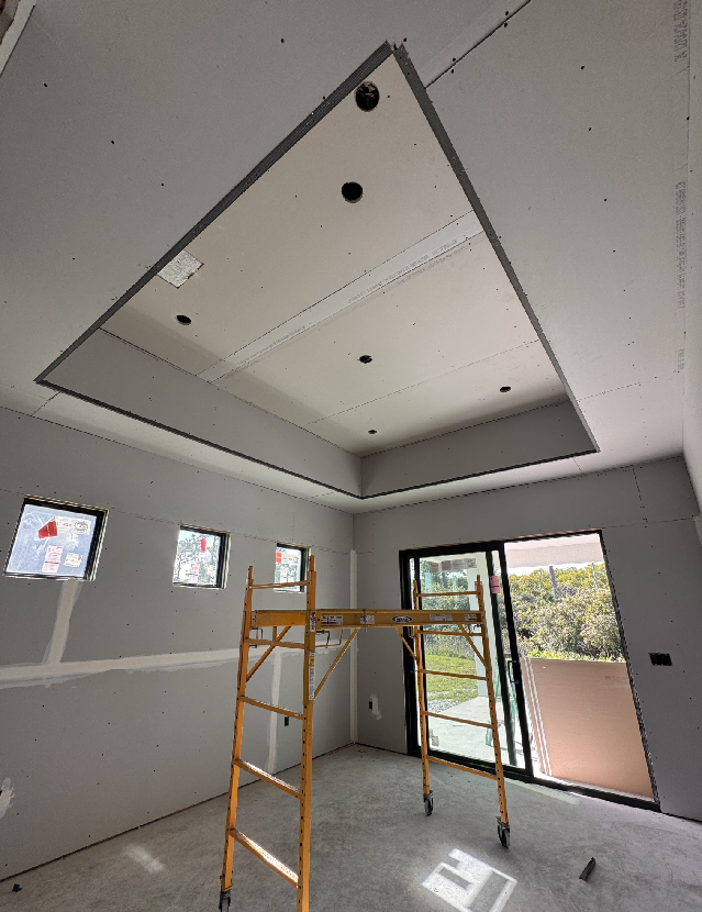 A room under construction with unfinished drywall and a large rectangular recess in the ceiling awaits the expertise of a general contractor. A yellow scaffold stands at the center, surrounded by three small square windows and a large glass door, where daylight streams through, illuminating the scene.