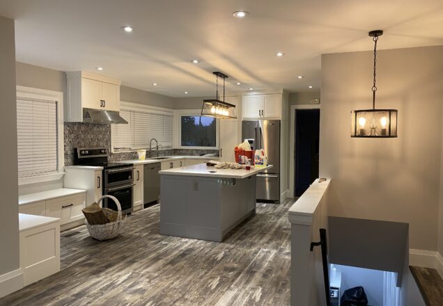Modern kitchen with wooden floors by a skilled flooring installation team, white cabinets, and a central island with pendant lights. Stainless steel appliances on the left, including an oven and fridge. A basket sits by the island. Stairs lead down on the right.