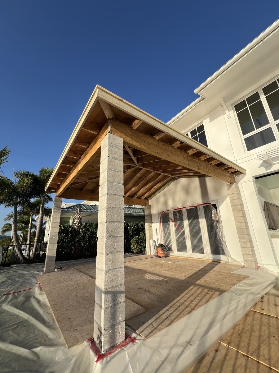 A partially constructed outdoor patio with a wooden roof supported by concrete pillars attached to a white house, showcasing building materials and tools—a perfect scene for any general contractor during a home renovation under a clear blue sky.