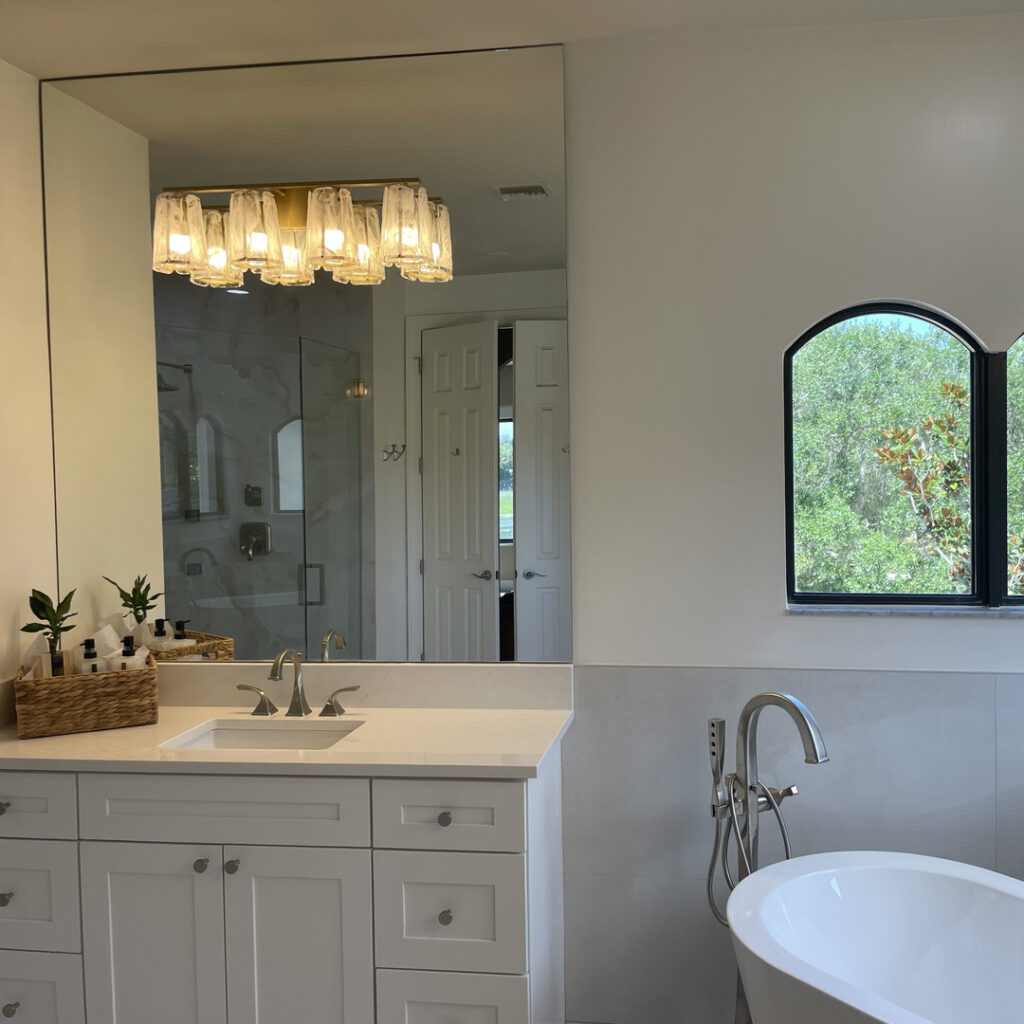 Modern bathroom with a white vanity, rectangular mirror, glass light fixture, potted plants, freestanding bathtub, and an arched window showing greenery—perfect inspiration for your next home renovation or to discuss with your general contractor.