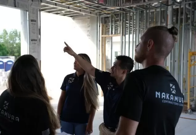 A group of four people wearing Nakamura Contractors shirts stands inside a building under construction. As a general contractor, one man points upward while the others look in the same direction, listening attentively.