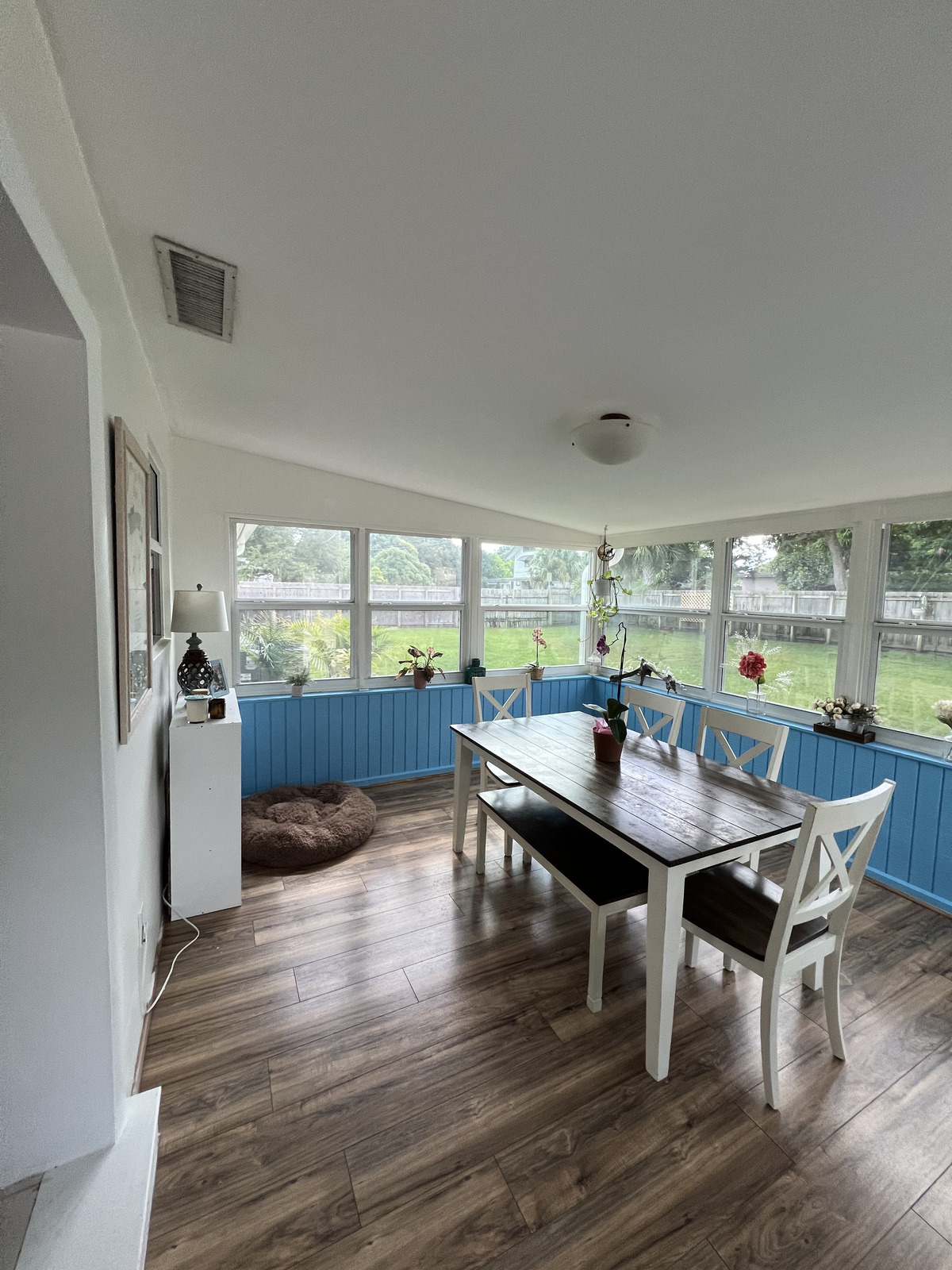 A bright sunroom with large windows, a wooden dining table, white chairs, potted plants, and a cozy dog bed on the floor—perfect inspiration for your next home renovation or to show your general contractor what you envision.