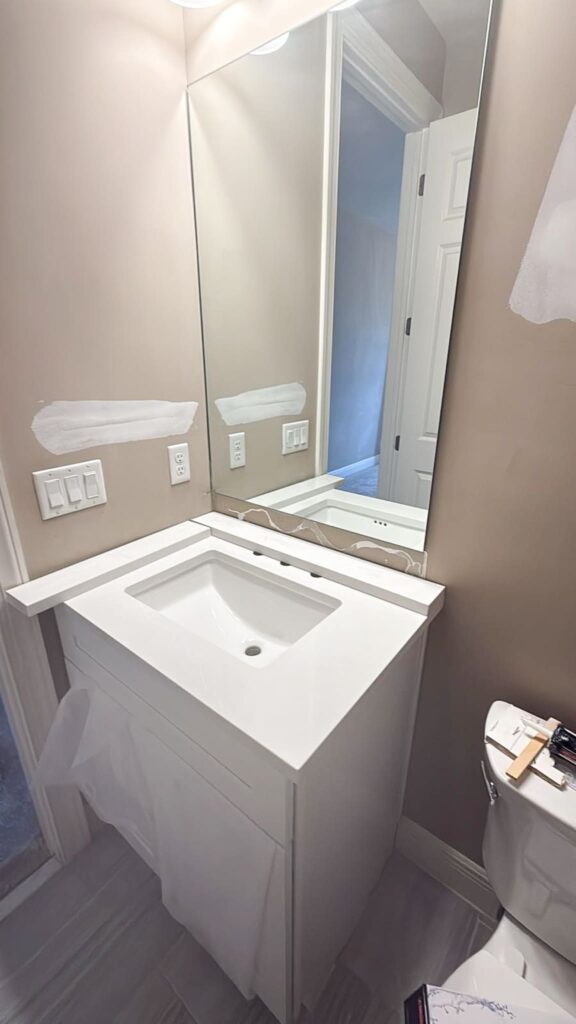 A modern bathroom with a large mirror, white sink vanity, light switches and outlet on the wall, beige walls with white paint patches, and part of a toilet visible in the lower right corner.
