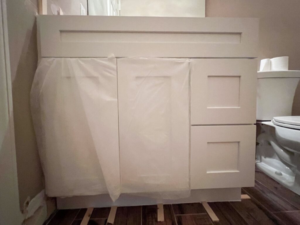 A white bathroom vanity with cabinet doors and drawers covered in protective plastic sits on a dark wood floor. Part of a toilet and toilet paper are visible to the right.