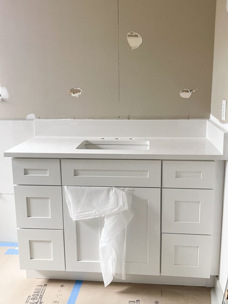 A white bathroom vanity with a sink is installed against an unfinished wall with exposed plumbing holes; a plastic sheet hangs from the sink area. The floor is covered for protection.