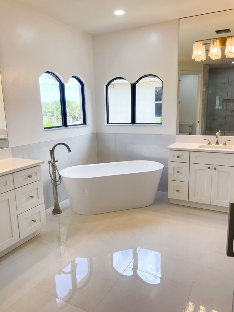 A modern bathroom with a freestanding white bathtub, two white vanities with sinks and cabinets, arched windows, large mirror, and glossy tiled floor. Natural light brightens the clean, minimalist space.