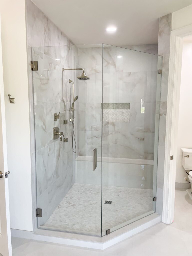 Modern bathroom shower with clear glass doors, marble-style tile walls, built-in shower bench, niche shelf, and stainless steel fixtures. A partial view of a toilet is visible outside the shower area.