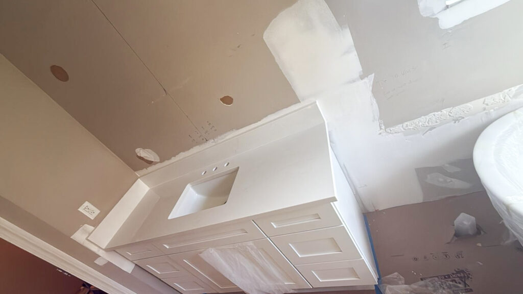 A bathroom under renovation with an unfinished wall, a white vanity with a sink, and protective plastic covering some surfaces. The walls show patches of drywall compound and unpainted sections.