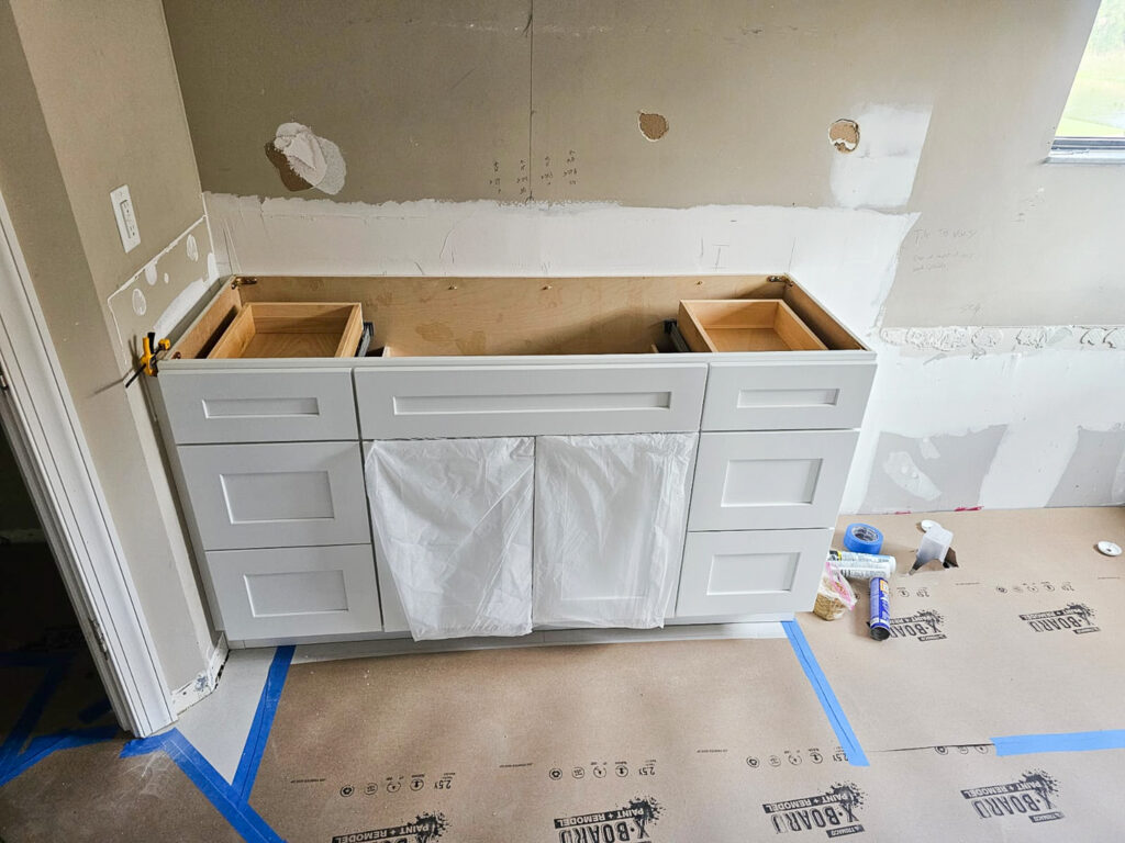 A white bathroom vanity without a countertop is installed in a partly renovated room. Two cabinet drawers are open, and plastic sheets hang on the front. Construction tools and supplies are scattered on the taped floor.