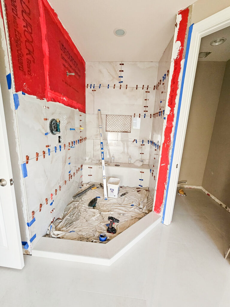 A partially tiled shower under construction, with tools, a bucket, and a tarp on the floor. Red waterproofing is visible on the walls, and tile spacers are holding tiles in place. An open door leads to another room.