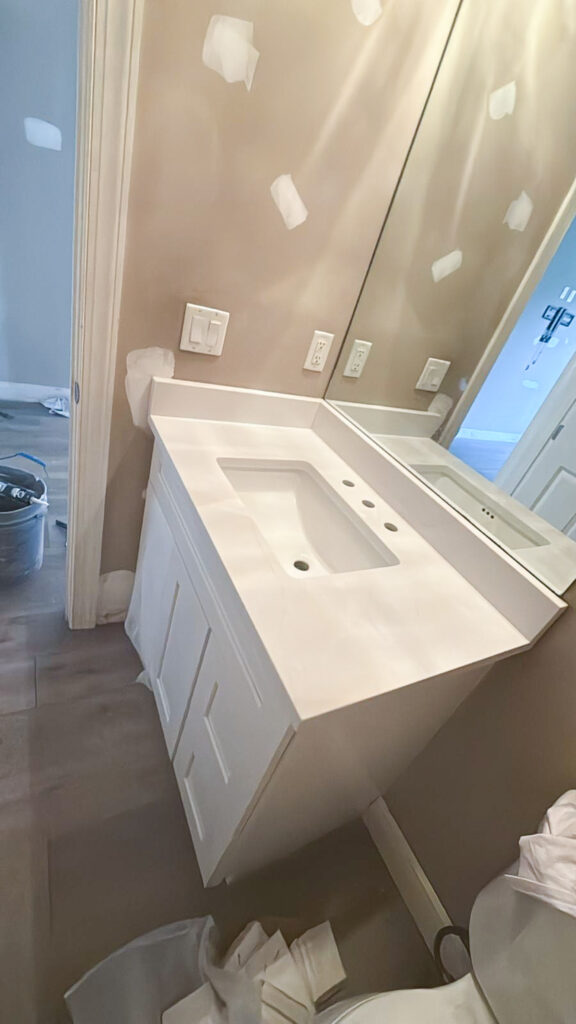 A bathroom under renovation with a white sink vanity, large mirror, and beige walls patched with white spackle. Construction materials and tools are scattered on the floor.