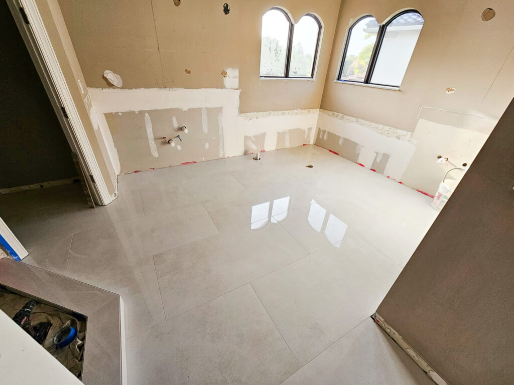 A room under renovation with new light-colored tile flooring, unfinished walls, exposed plumbing, and two arched windows letting in natural light. Construction materials are present in the space.