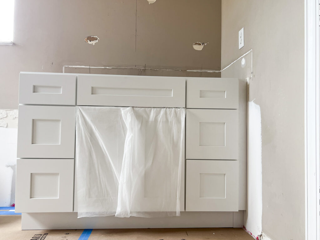 White bathroom vanity with shaker-style doors and drawers is installed against a beige wall. A plastic sheet covers the middle area, and unfinished wall sections and exposed outlets are visible, indicating ongoing renovation.