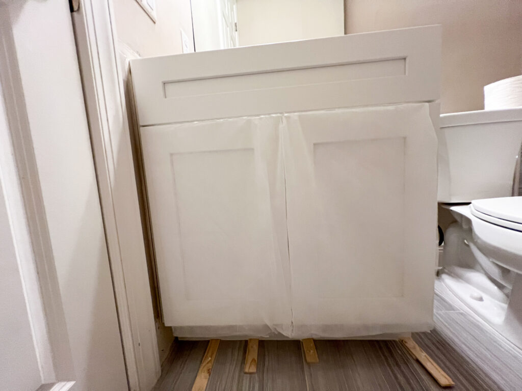 A white bathroom vanity cabinet with protective plastic wrapping stands on wooden spacers near a toilet, against a beige wall and gray tile floor.