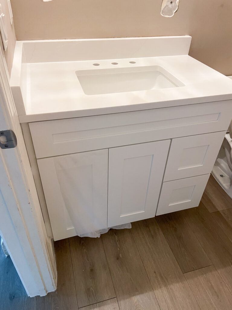 A white bathroom vanity with a built-in sink, set against a beige wall with unfinished patches. The vanity has two cabinet doors, three drawers, and sits on light wood flooring. A plastic sheet is partially visible under the cabinet.