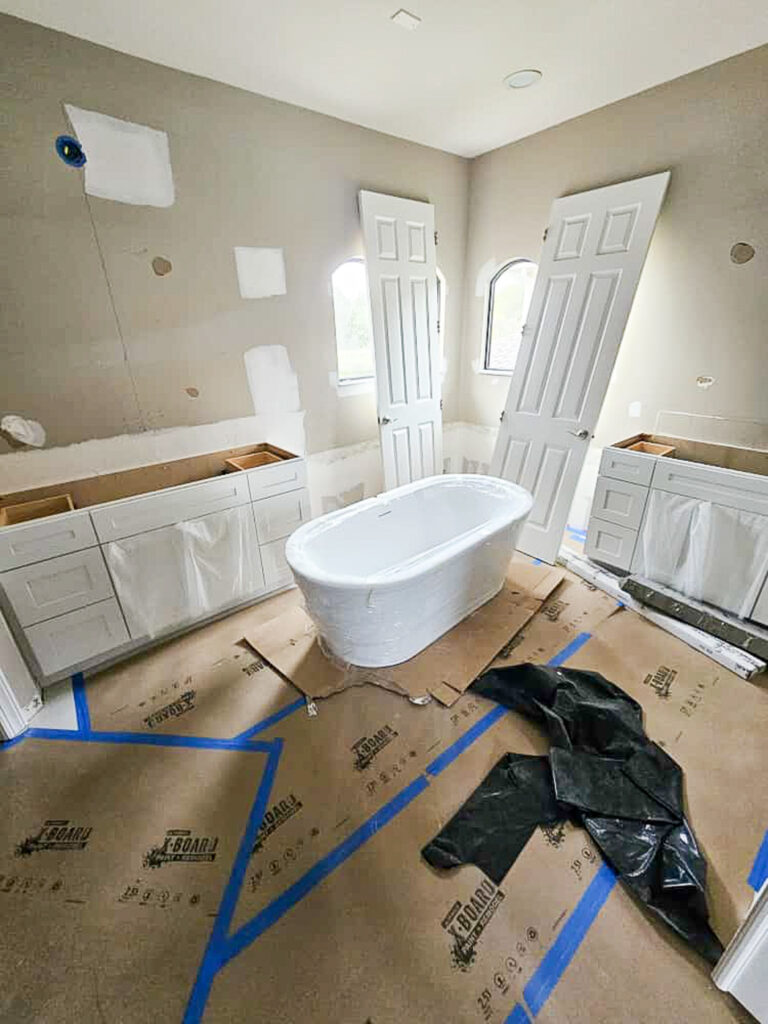A bathroom under construction with a freestanding bathtub wrapped in plastic, two vanities without countertops, unpainted drywall patches, and protective covering on the floor. Two doors lean against the wall.