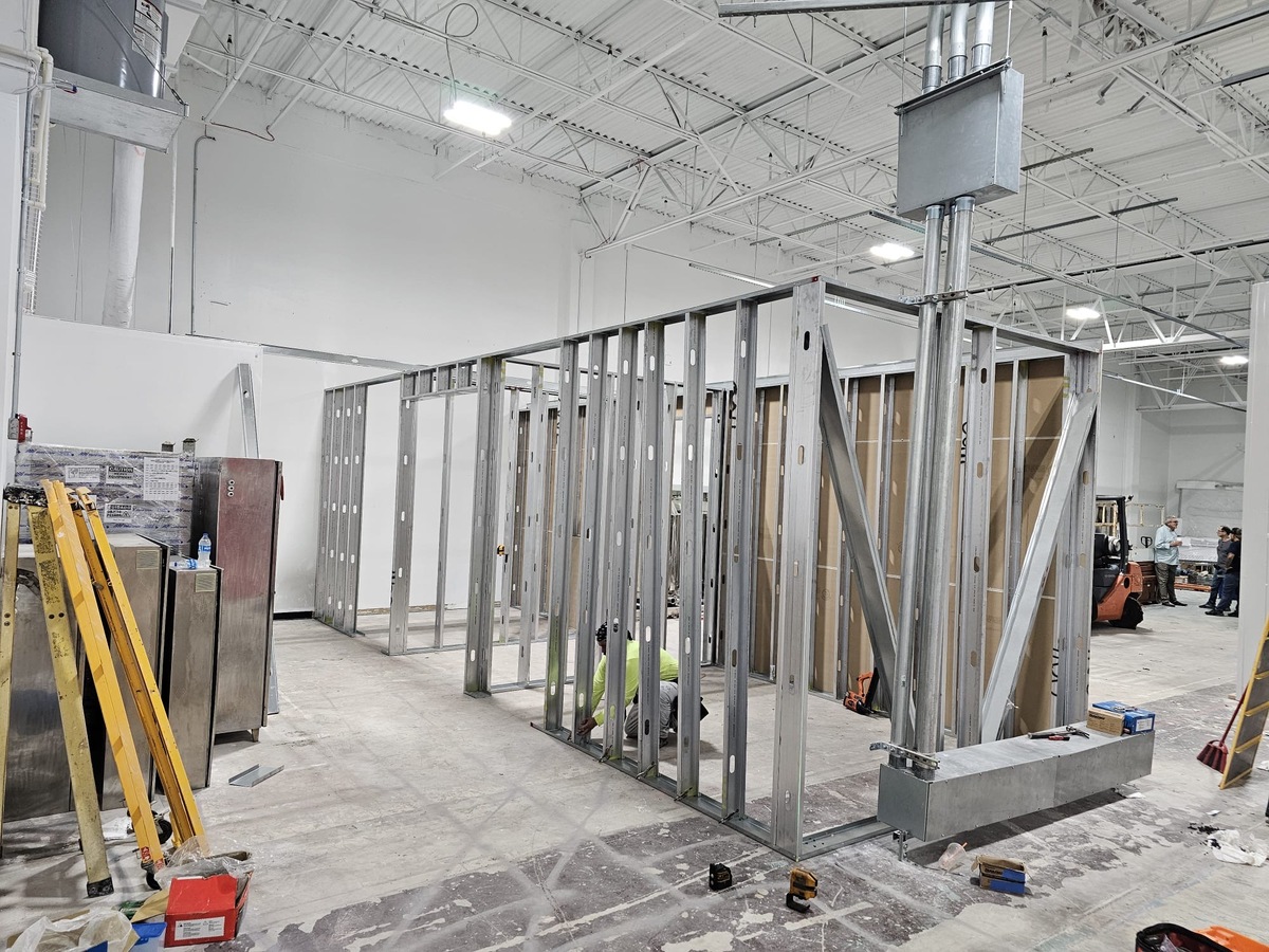 A construction worker, hired by a general contractor, installs metal wall studs inside a large industrial building with white walls, high ceilings, and exposed pipes. Tools, ladders, and construction materials are scattered on the floor.