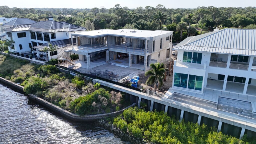 A waterfront modern house under construction, managed by a general contractor, is surrounded by greenery with neighboring finished homes and a water view. Construction materials and a blue dumpster are visible on site.