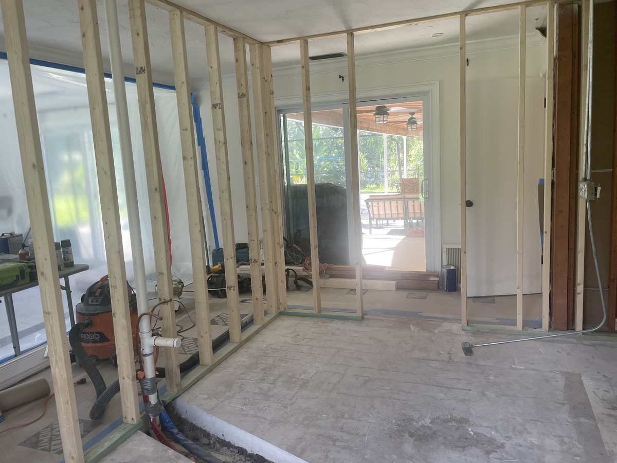 A room under home renovation with exposed wooden wall studs, construction tools, and piping on the floor. A sliding glass door opens to a sunroom with outdoor seating and greenery visible through the windows.