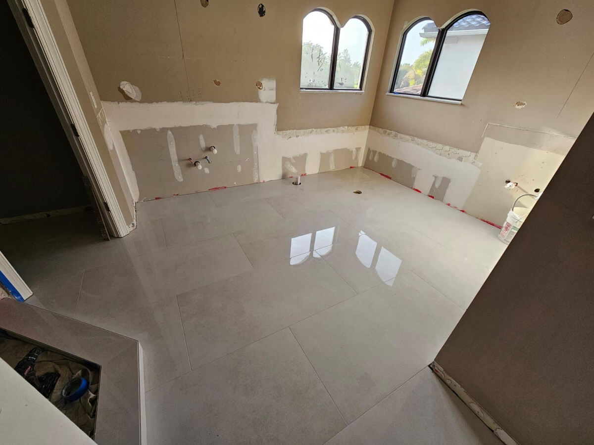 A partially renovated bathroom by a skilled general contractor features newly installed large, glossy floor tiles. The walls, still unfinished with exposed plumbing and no fixtures, highlight the home renovation process as light pours in through three arched windows.