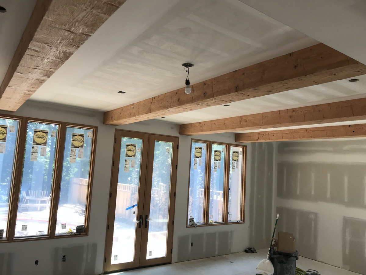 The interior of a room under construction showcases unfinished drywall, exposed wooden ceiling beams, a bare light bulb, and large windows with French doors—a typical scene during home renovation managed by a general contractor. Construction materials are scattered on the floor.