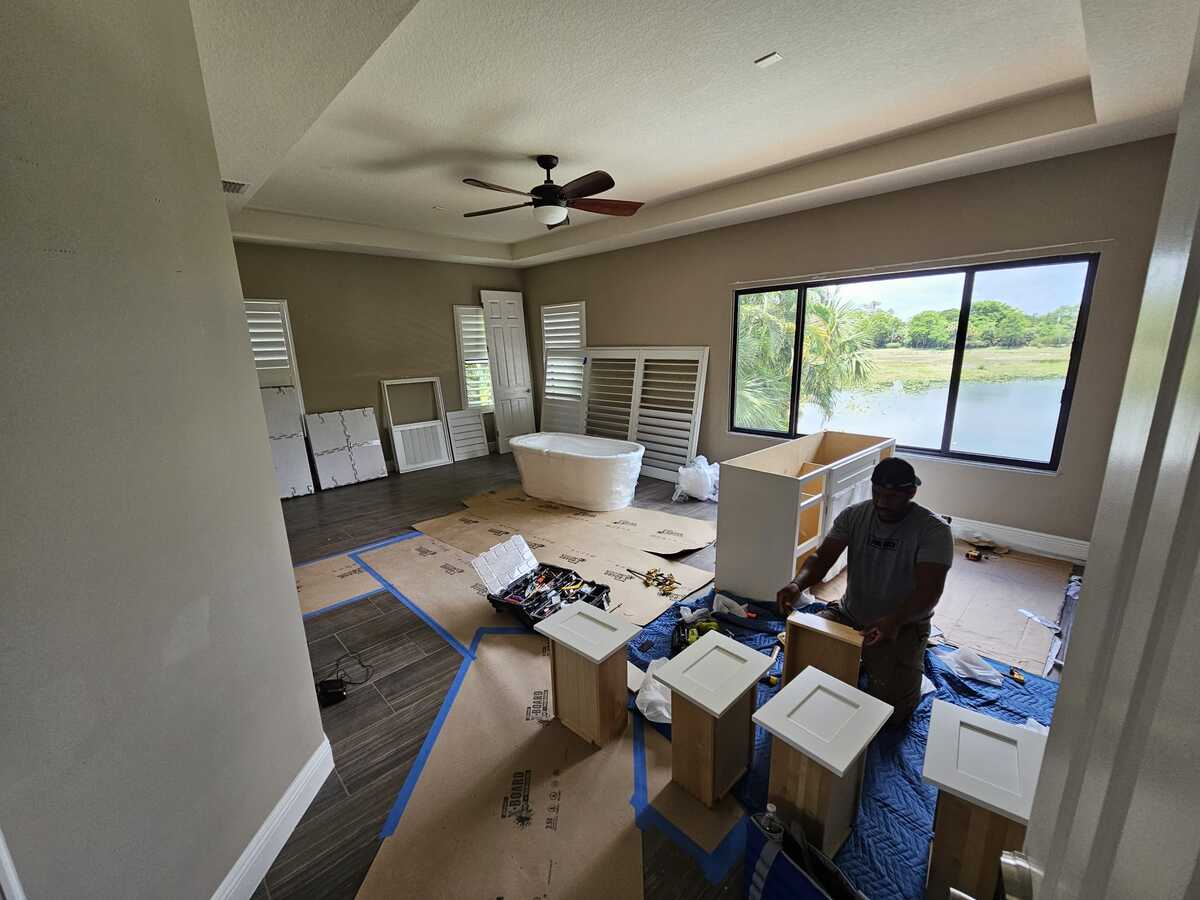 A person works on assembling bathroom cabinets as part of a home renovation in a large, bright room with a ceiling fan, freestanding bathtub, and big windows overlooking greenery and water. Tools and materials are spread on the floor.