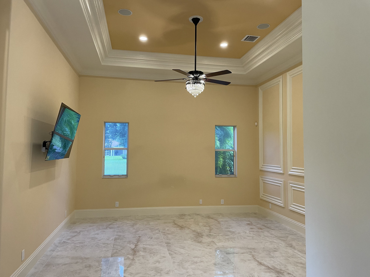 A spacious, empty room with beige walls, white trim, marble tile floor, two small windows, and a ceiling fan with a light fixture—perfect for your next home renovation or as envisioned by a skilled general contractor. A wall-mounted TV displays a nature scene.