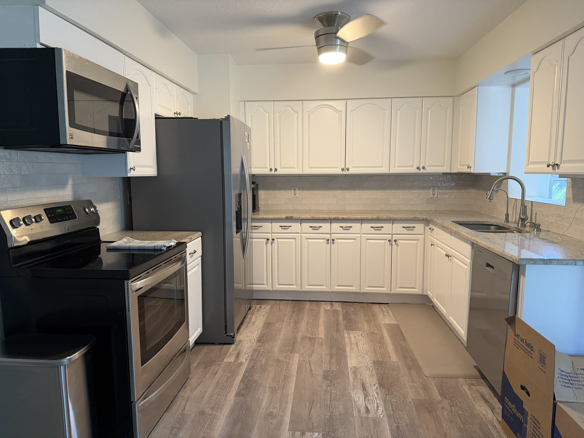 Modern kitchen with white cabinets and stainless steel appliances, perfect for your next home renovation. Features a gray refrigerator, electric stove, built-in microwave, window-side sink, wood-look flooring, ceiling fan, and a cardboard box on the right.