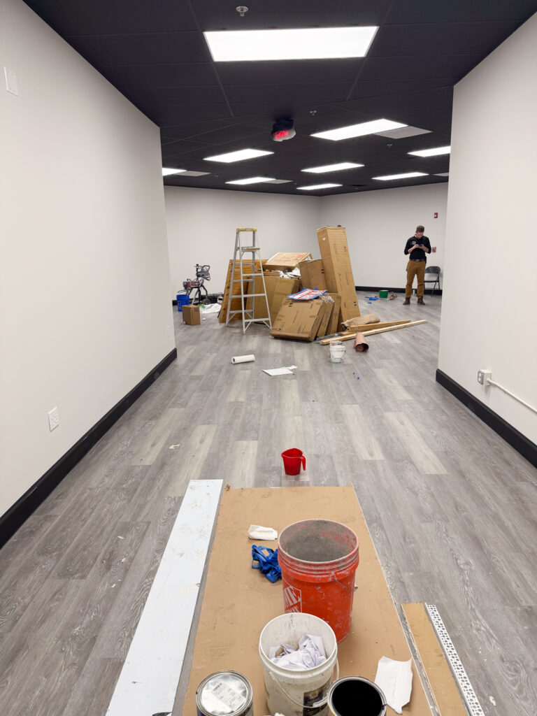 A room under renovation with scattered tools, paint cans, a red bucket, cardboard boxes, a stepladder, and two people working in the background. The floor is wood-look, and the ceiling has bright lights.