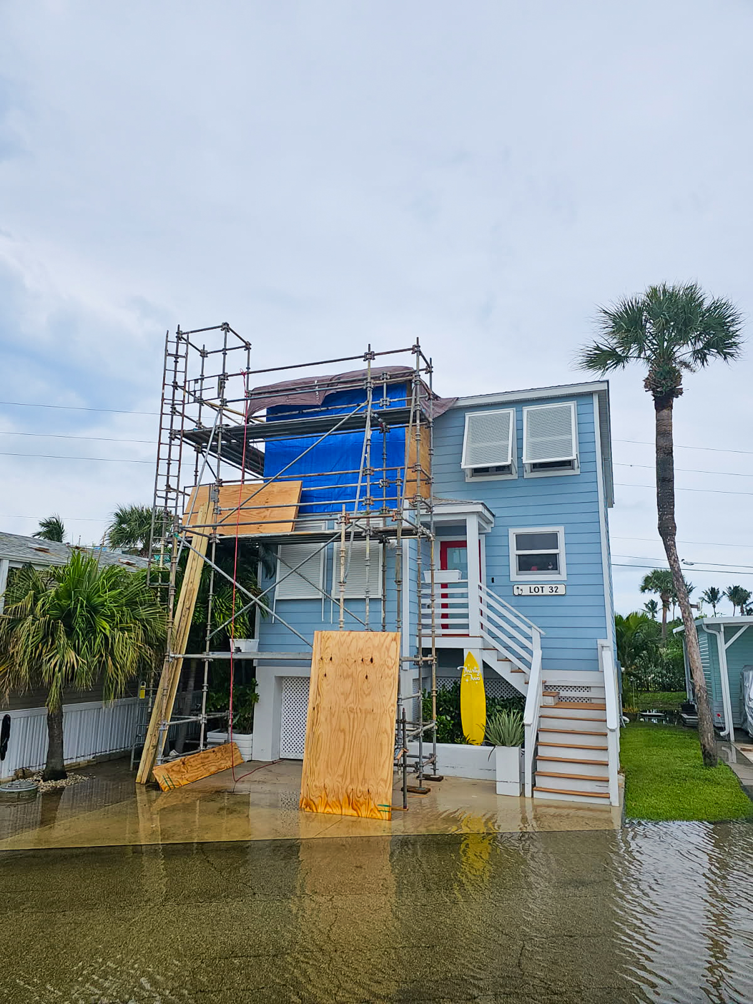 A blue house on stilts with scaffolding and plywood covering part of the upper floor. A yellow surfboard leans near the stairs. Flood water covers the ground, and palm trees are visible nearby.