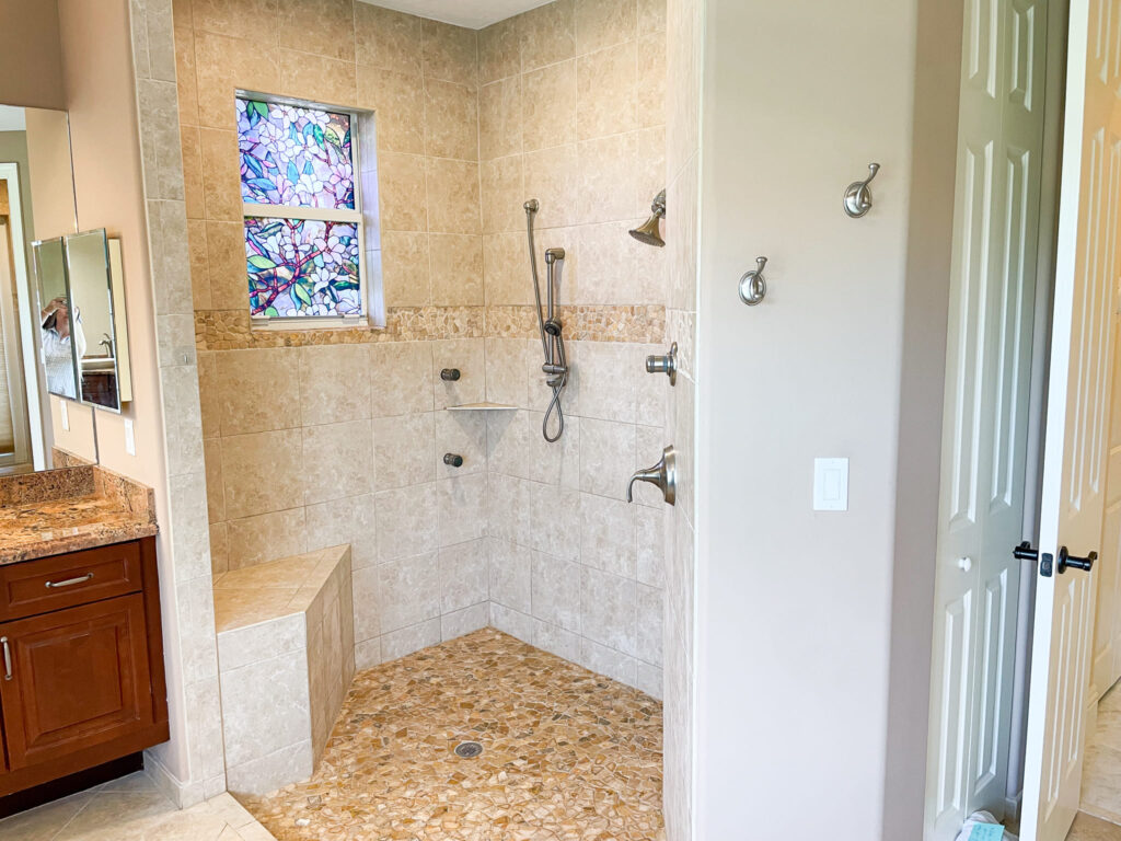 Spacious walk-in shower with beige tile, built-in bench, rainfall showerhead, hand-held shower, and grab bars. A stained glass window adds color. Nearby, there are three wall hooks and a vanity with a granite countertop.