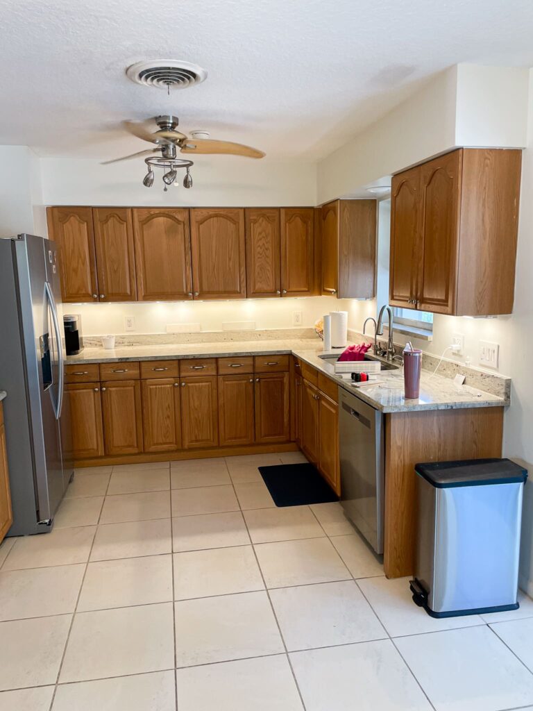 A clean kitchen with wooden cabinets, stainless steel appliances, white tile floor, granite countertops, a ceiling fan, and a trash bin. A red dish rack and a tumbler sit near the sink.