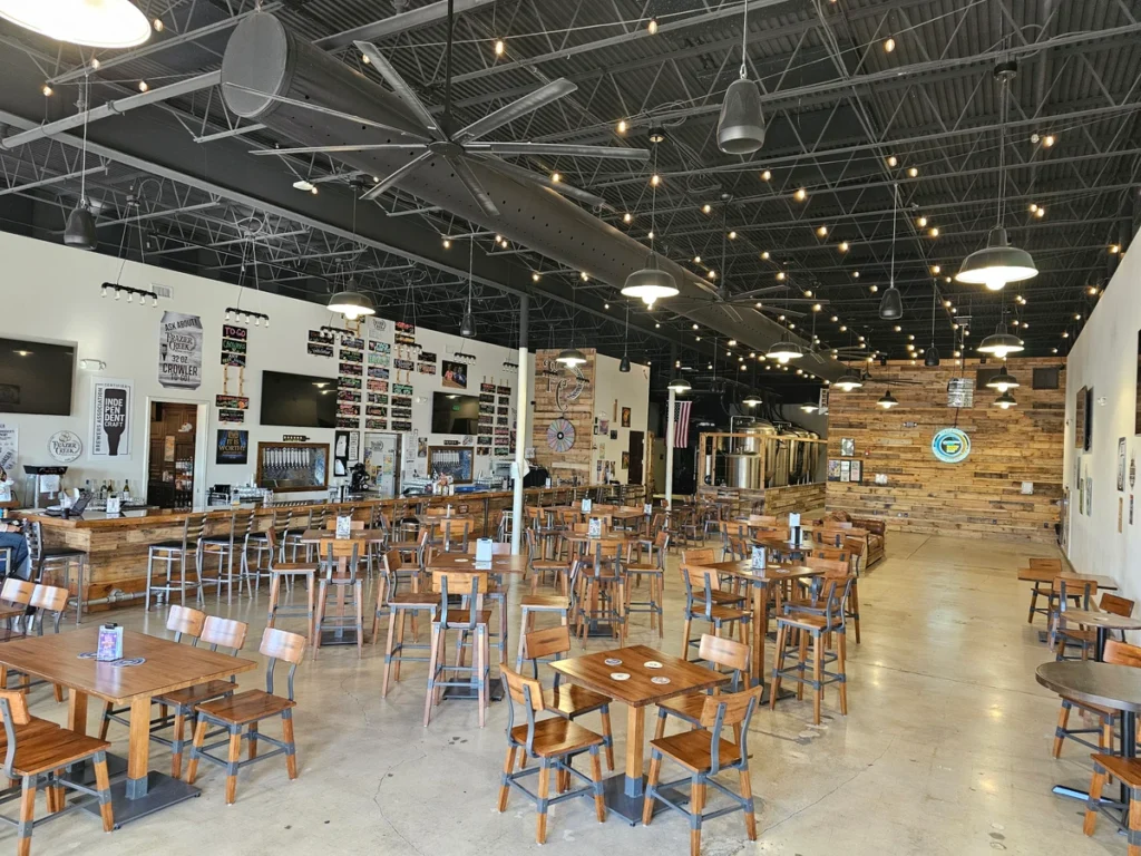 Spacious brewery taproom with wooden tables and chairs, high ceilings, string lights, a long bar on the left, wall menus with beer options, and brewing tanks in the back. The space is empty of people.
