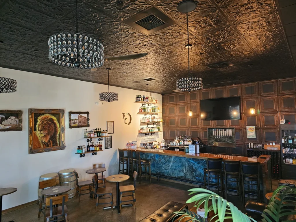 A cozy bar with a dark ornate ceiling, crystal chandeliers, a lion painting on the wall, a stocked shelf of bottles behind the bar, high chairs at the counter, and small round tables with wooden stools.