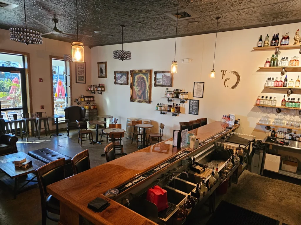 A cozy café with wooden tables and chairs, a long bar counter, shelves of bottles on the wall, warm lighting, art on the walls, and large windows letting in natural light.