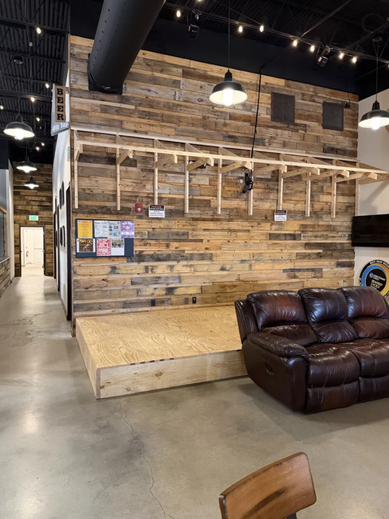 A seating area with a brown leather couch on a wooden platform, set against a rustic wooden wall with exposed beams. Overhead lights hang from a black ceiling, and a hallway is visible to the left.
