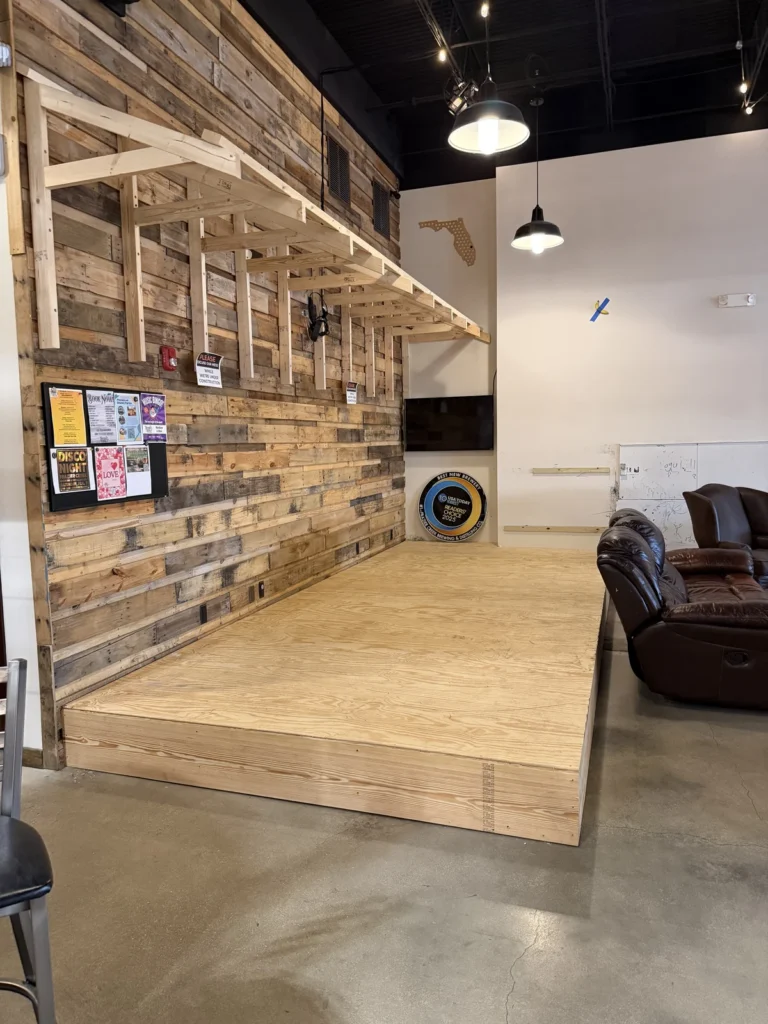 A small wooden stage with a rustic wood-paneled wall, bulletin board with flyers, and exposed beams. A brown leather couch sits beside the stage on a polished concrete floor under hanging lights.