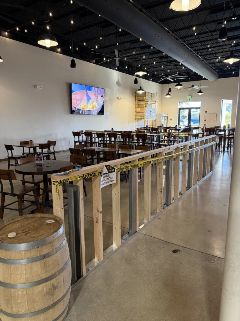 A restaurant dining area with empty wooden tables and chairs, a construction barrier with caution tape, a barrel, and a TV on the wall. Bright lighting and visible ceiling fixtures.