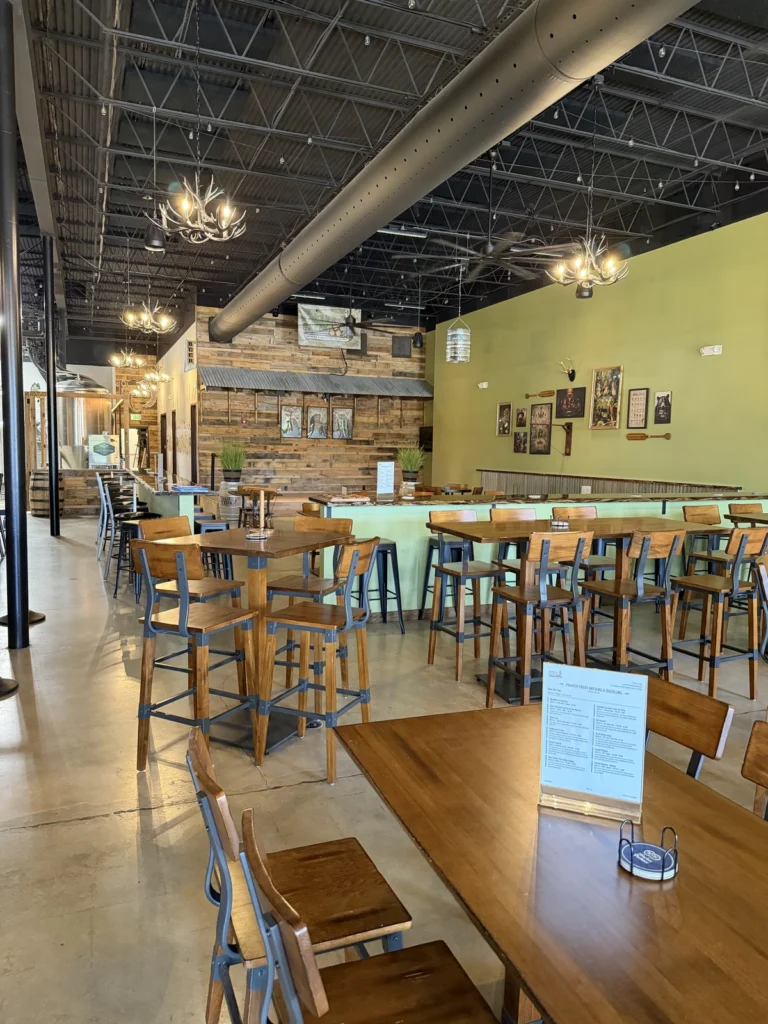 A spacious, modern restaurant with high ceilings, wooden tables and chairs, green accent walls, rustic decor, and framed pictures on the wall; ceiling lights and natural light brighten the space.