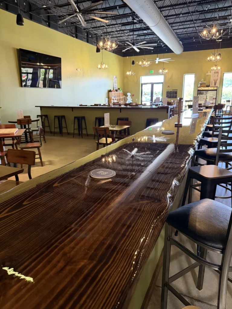 A modern, well-lit bar with a long, glossy wooden counter, high chairs, and tables. The space features light green walls, a mounted TV, hanging lights, and large windows letting in natural light.