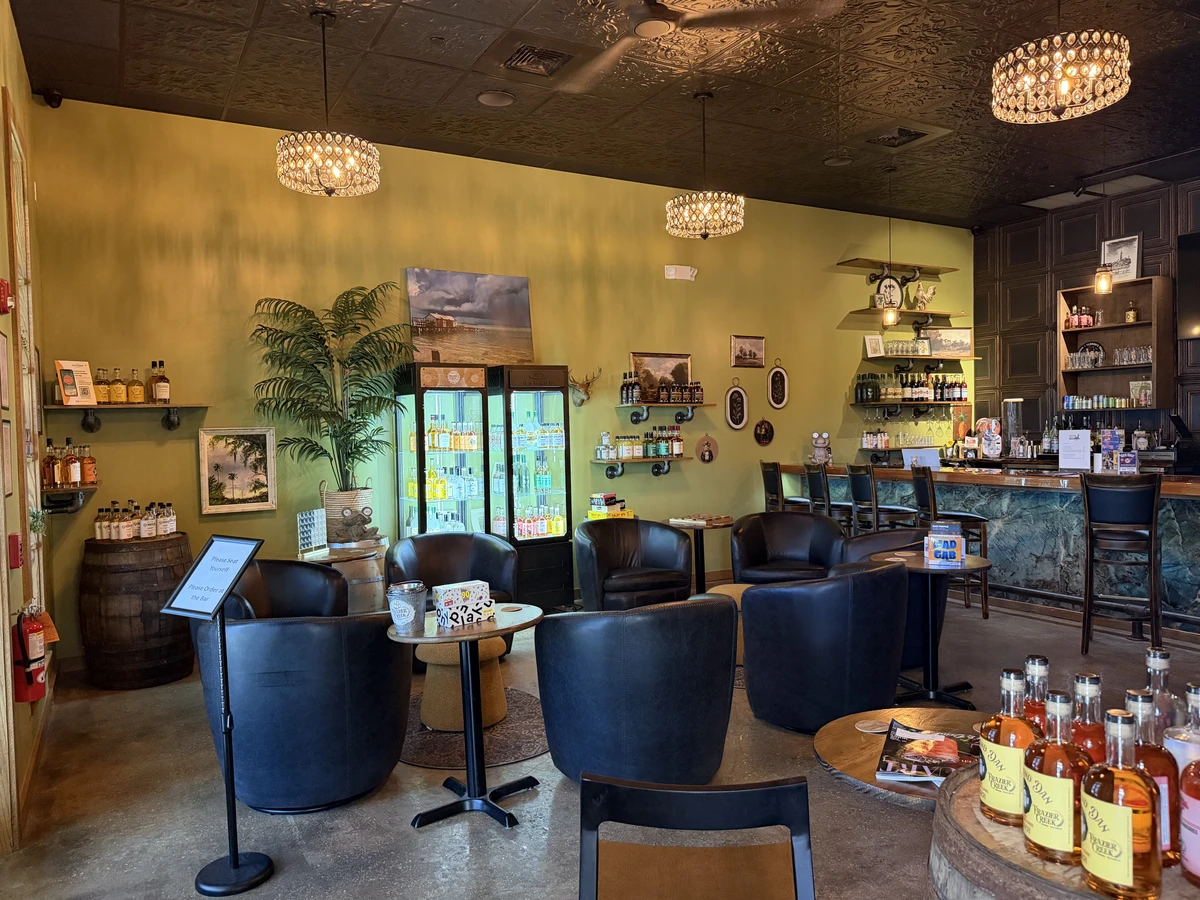 A cozy café interior with black leather chairs, round tables, a bar area, a fridge stocked with drinks, and shelves displaying bottles and décor against yellow-green walls under hanging chandeliers.