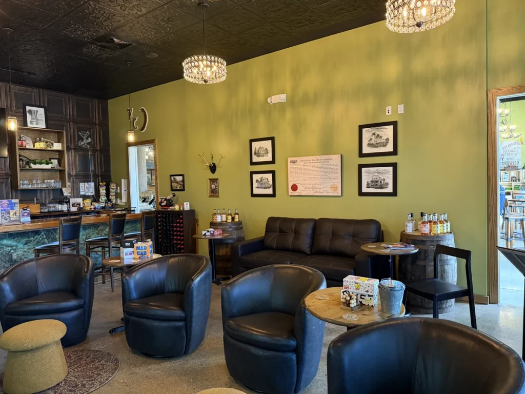 Cozy cafe interior with black leather chairs, round wooden tables, a brown couch, and framed art on a green wall. A bar area with stools and shelves is in the background, and two chandeliers hang from the ceiling.